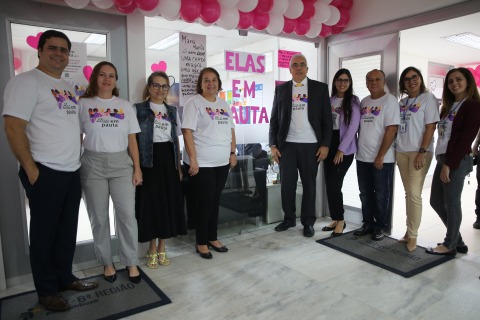 Pessoas usando a mesma camisa posando junto a uma porta de vidro onde está escrito Elas em Pauta